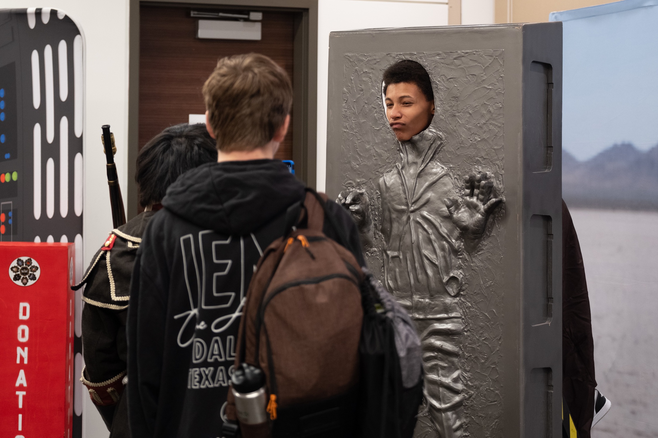 A fan poses as Han Solo frozen in carbonite, sticking his head through the display while other attendees look on and laugh.