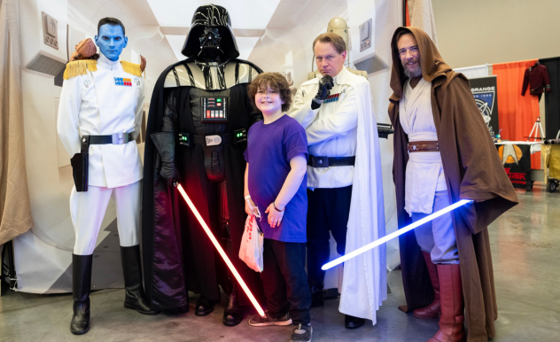 The image features a young fan posing with four individuals dressed as characters from the "Star Wars" universe at a FAN EXPO event. The characters include Grand Admiral Thrawn, Darth Vader, Grand Moff Tarkin, and Obi-Wan Kenobi, each in highly detailed costumes complete with iconic lightsabers and military regalia. The child, standing in the center, smiles broadly, clearly thrilled to be part of the scene. This moment captures the excitement and engagement of younger attendees with their favorite characters, highlighting the immersive and fan-friendly atmosphere of the expo.