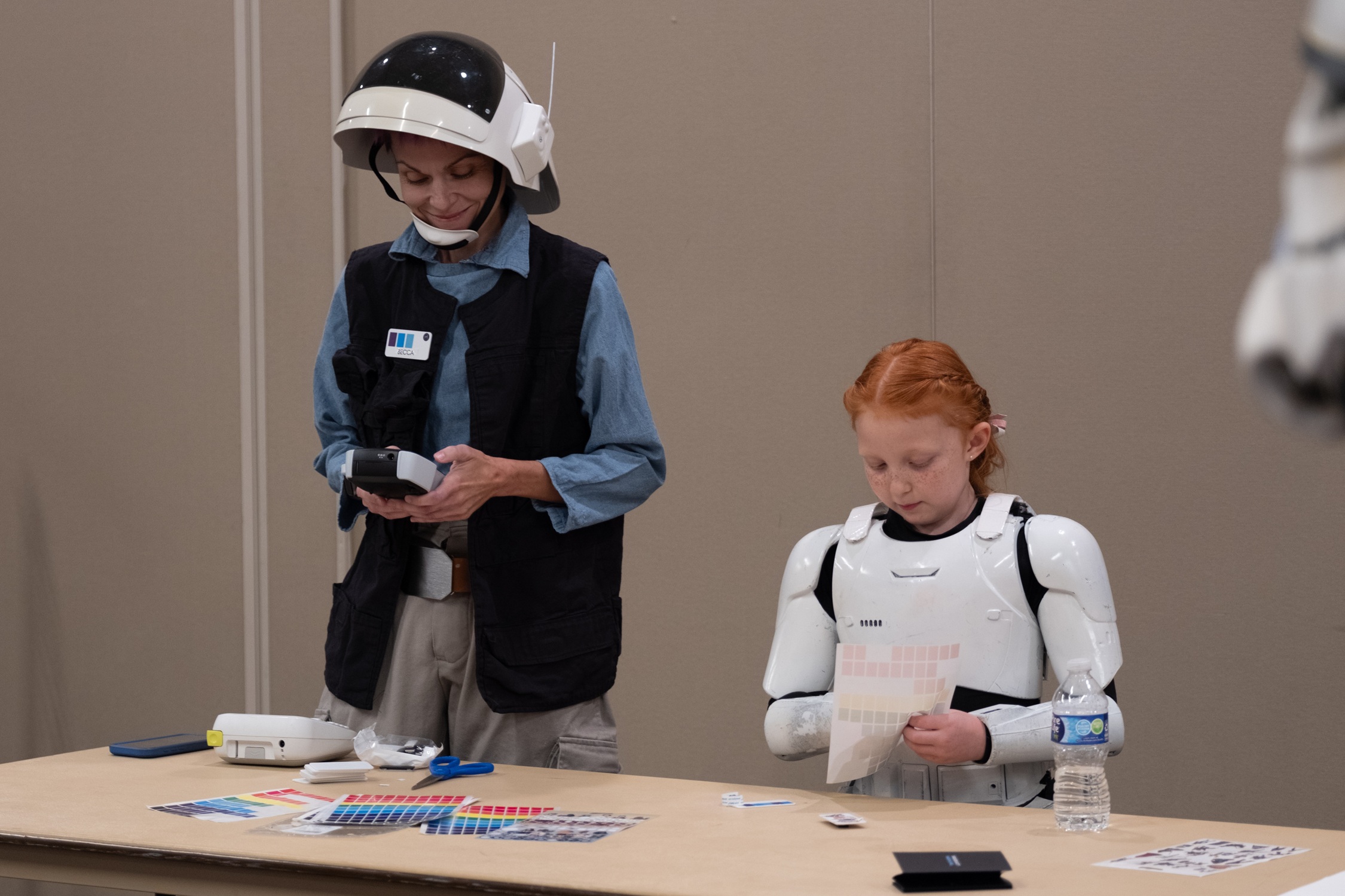 A woman dressed as a Star Wars Rebel officer and a young girl in Stormtrooper armor work on a craft activity together at a table.