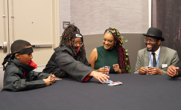 The image depicts a joyful moment at a FAN EXPO event, where a family is engaged in a card game. Two children and two adults, all dressed in costumes, are gathered around a table. The boy on the left, dressed in a ninja-themed outfit, is playfully reaching across the table to grab a card. The woman in the center, adorned with green and red braided hair, laughs along with the man on the right, who is wearing a formal suit and hat, suggesting a playful and light-hearted interaction. This scene captures a slice of family fun at the event, highlighting the inclusive and entertaining atmosphere that such conventions offer to attendees of all ages.