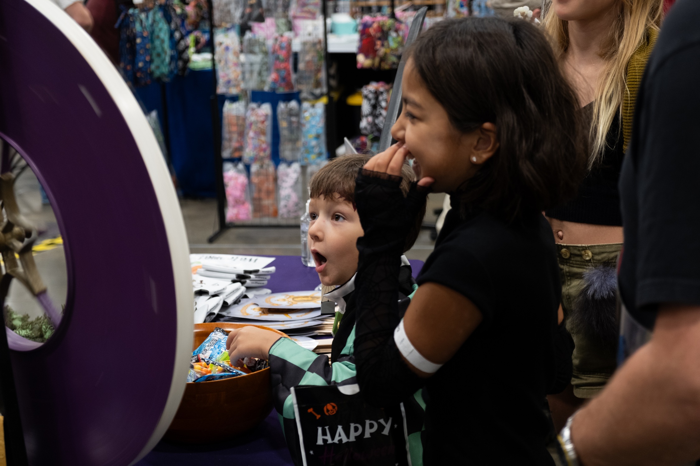 Two children smile excitedly while playing a prize wheel game at a booth, surrounded by candy and Halloween-themed giveaways.