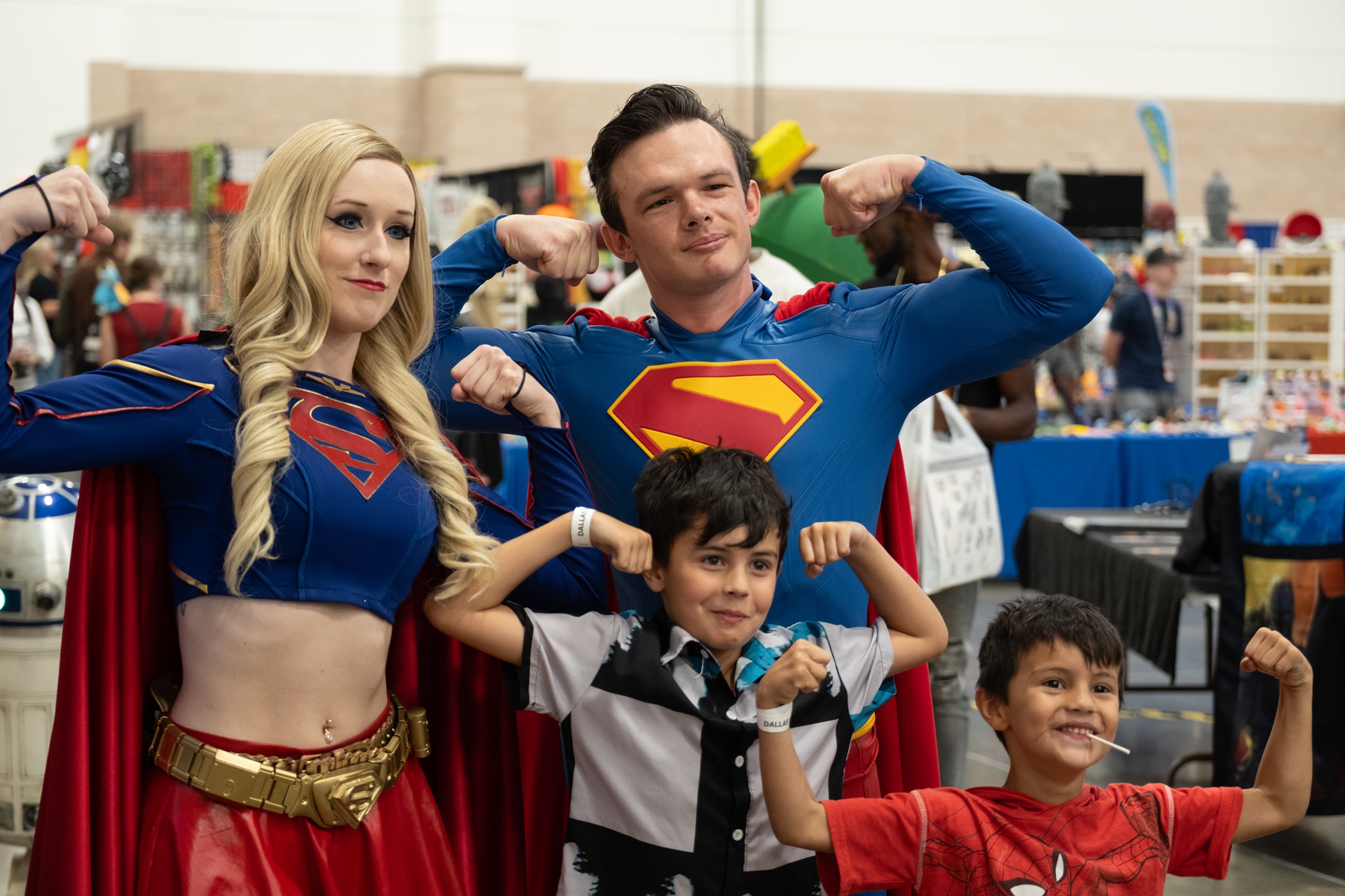 Cosplayers dressed as Supergirl and Superman flex their muscles alongside two young fans posing proudly in superhero stances.