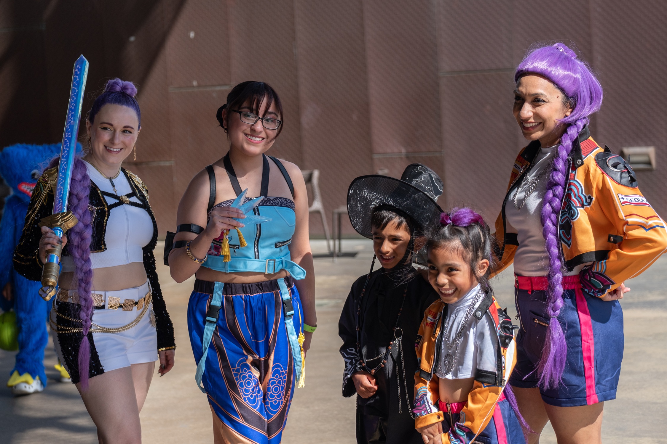 A group of cosplayers pose together outside, including two women and two children dressed in colorful, futuristic outfits inspired by anime or gaming characters.