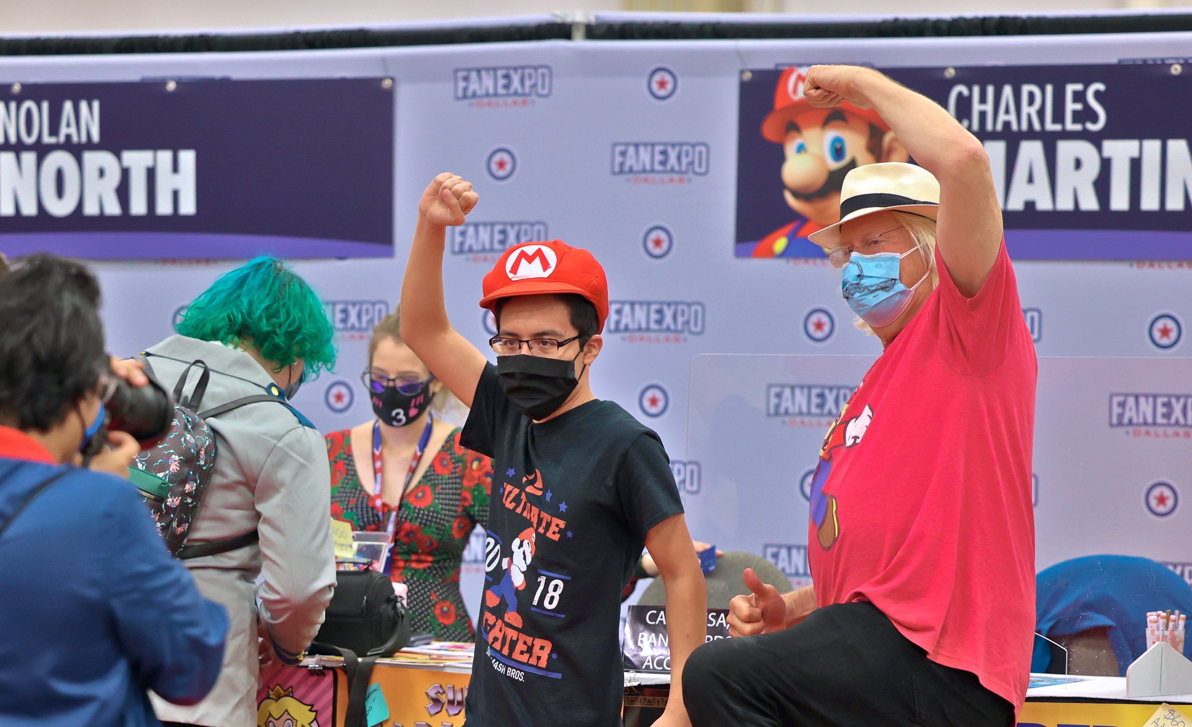 Charles Martinet in a white fedora and a red Mario shirt. He is posing in the Mario jump pose in front of his autograph table with a fan dressed in Mario shirt and hat.