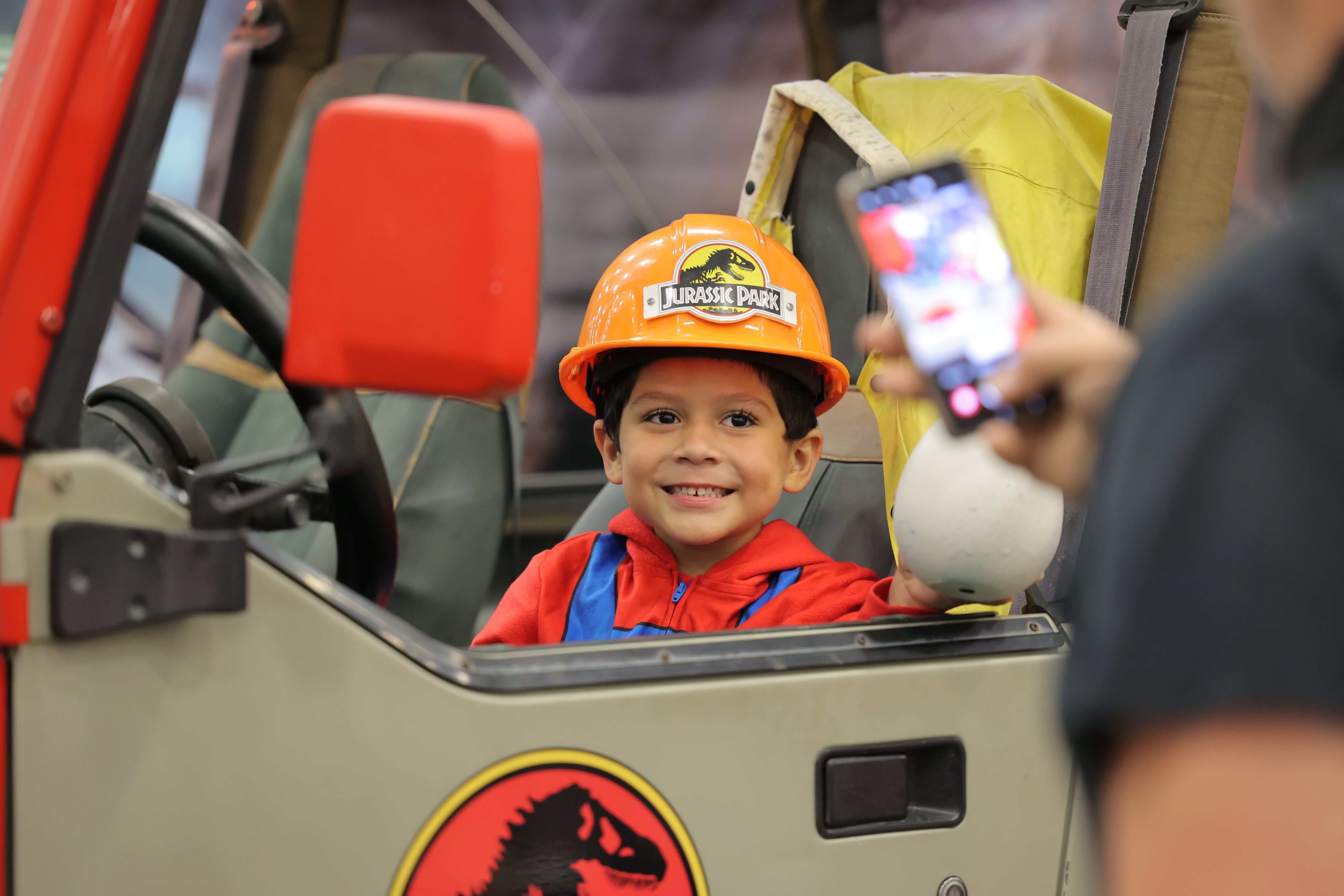 A child in a Jurrasic Park hard hat poses for a phone photo in a Jurrasic Park jeep
