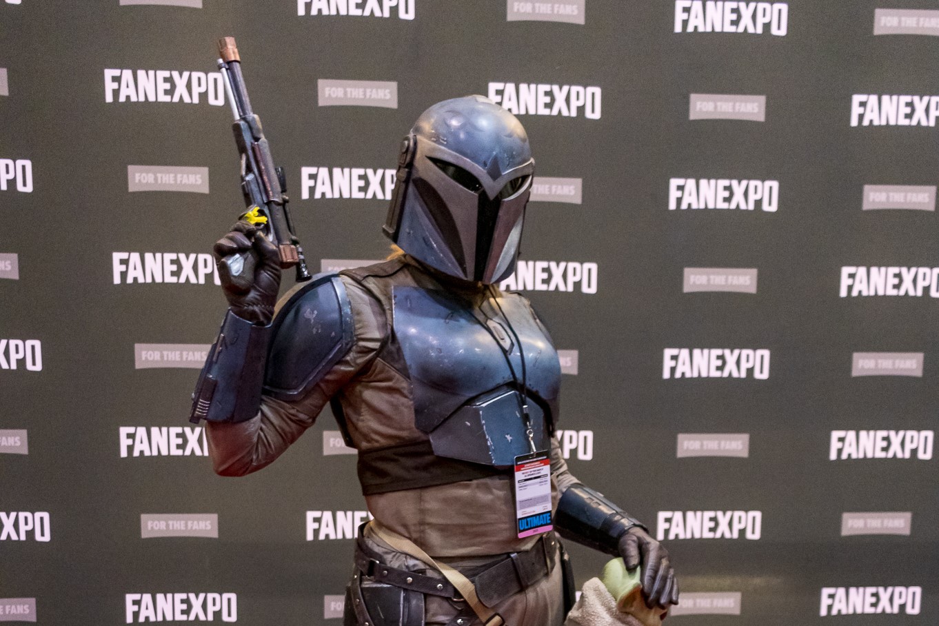 A cosplayer dressed as Bo-Katan Kryze from The Clone Wars & The Mandalorian, posing with her blaster as she pets Grogu.