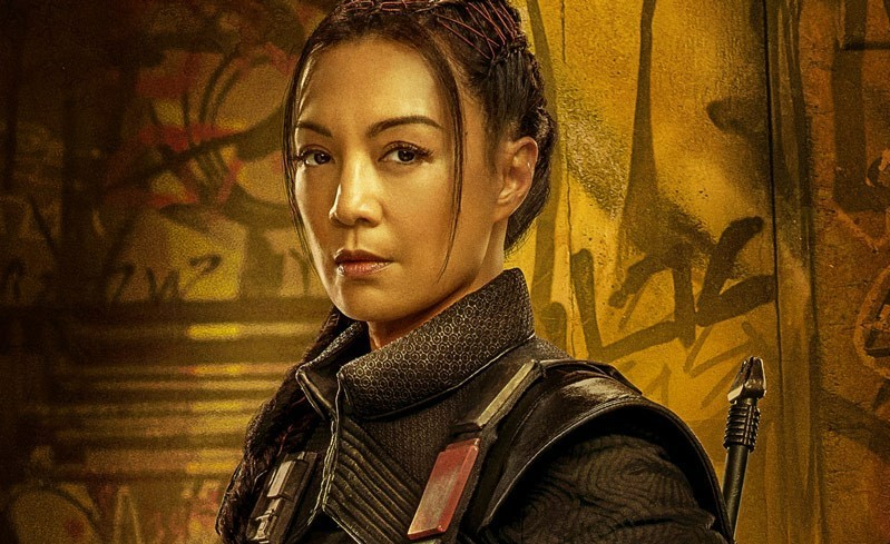 Ming-Na Wen as Fennec Shand. She is looking at the camera, 3/4 turn toward center. She is wearing a vest and strapped to her back is the end of a laser rifle. In the background there is alien writing on a yellowed wall.