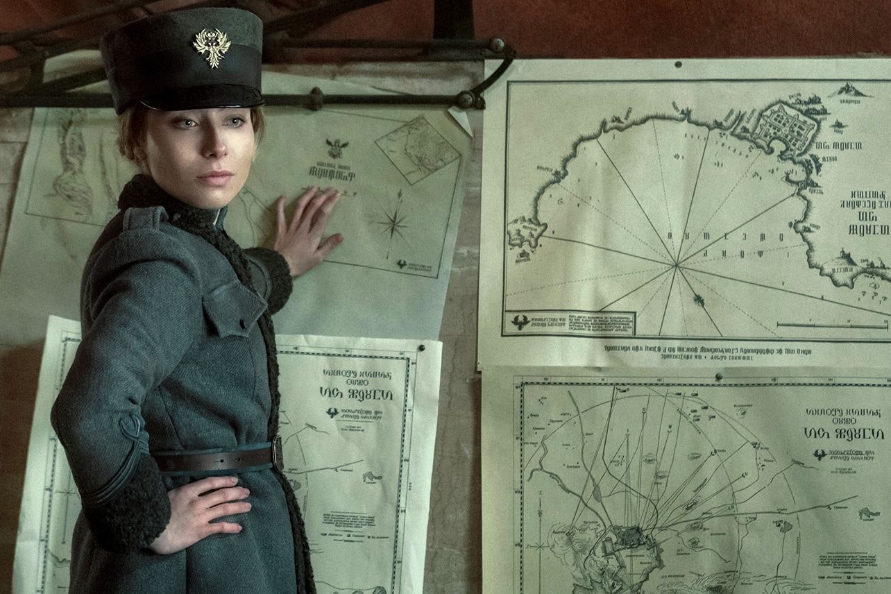 A character in a grey jacket and grey hat puts her hand on a series of old maps and charts