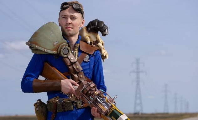 A man in a blue outfit with leather straps across holds a large prop weapon on an empty street
