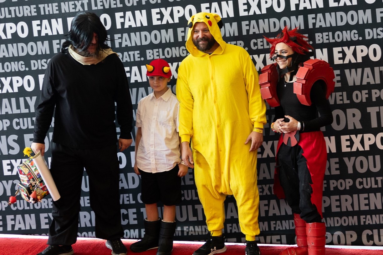 A group of cosplayers dressed as characters from various anime and pop culture series, including one in a Pikachu onesie, standing in front of a FAN EXPO backdrop.