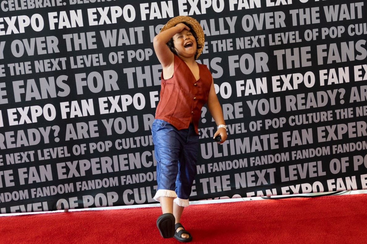 A young attendee dressed as Luffy from One Piece, striking a playful pose with a wide smile in front of a FAN EXPO backdrop.