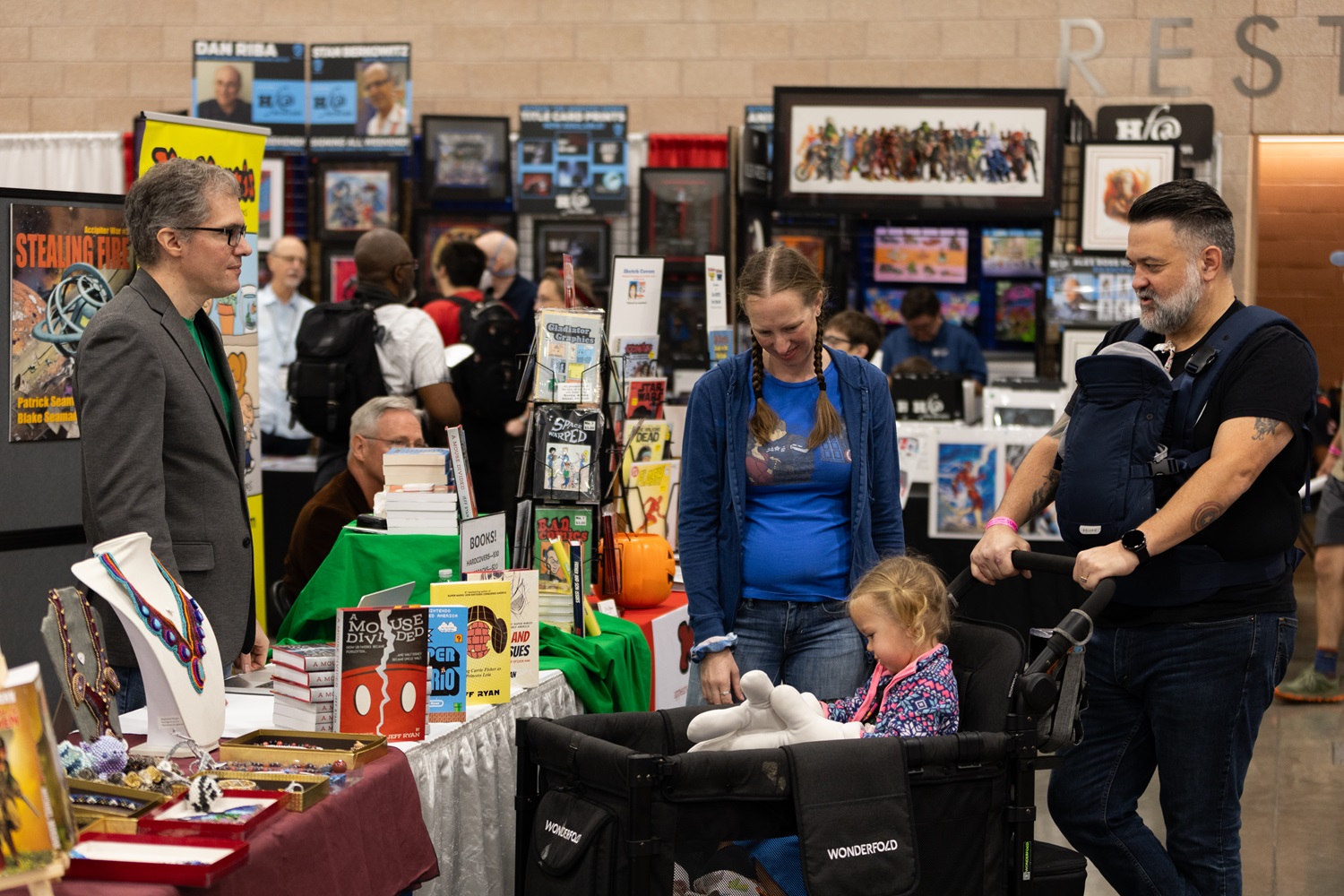 A family with two young children interacting with a vendor at a comic book and collectibles booth, surrounded by colorful books and merchandise at FAN EXPO.