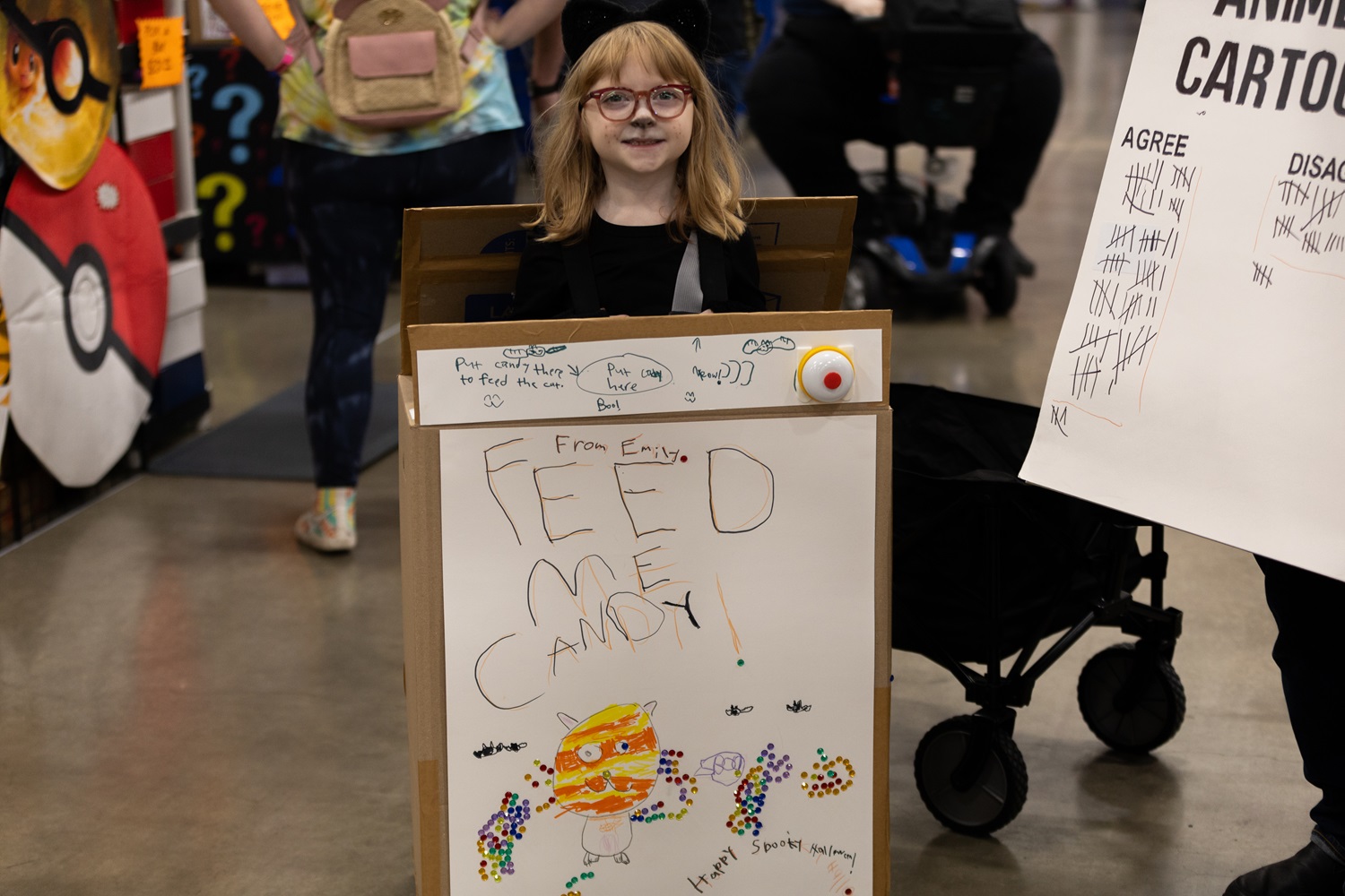A young girl dressed as a candy vending machine, complete with a hand-drawn “Feed Me Candy” sign, smiling at FAN EXPO.
