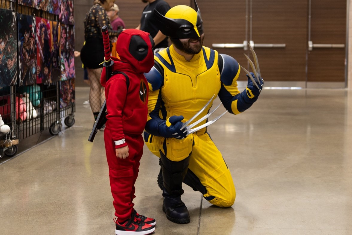 A child dressed as Deadpool standing next to an adult cosplayer dressed as Wolverine, both in colorful costumes, posing together on the show floor at FAN EXPO.
