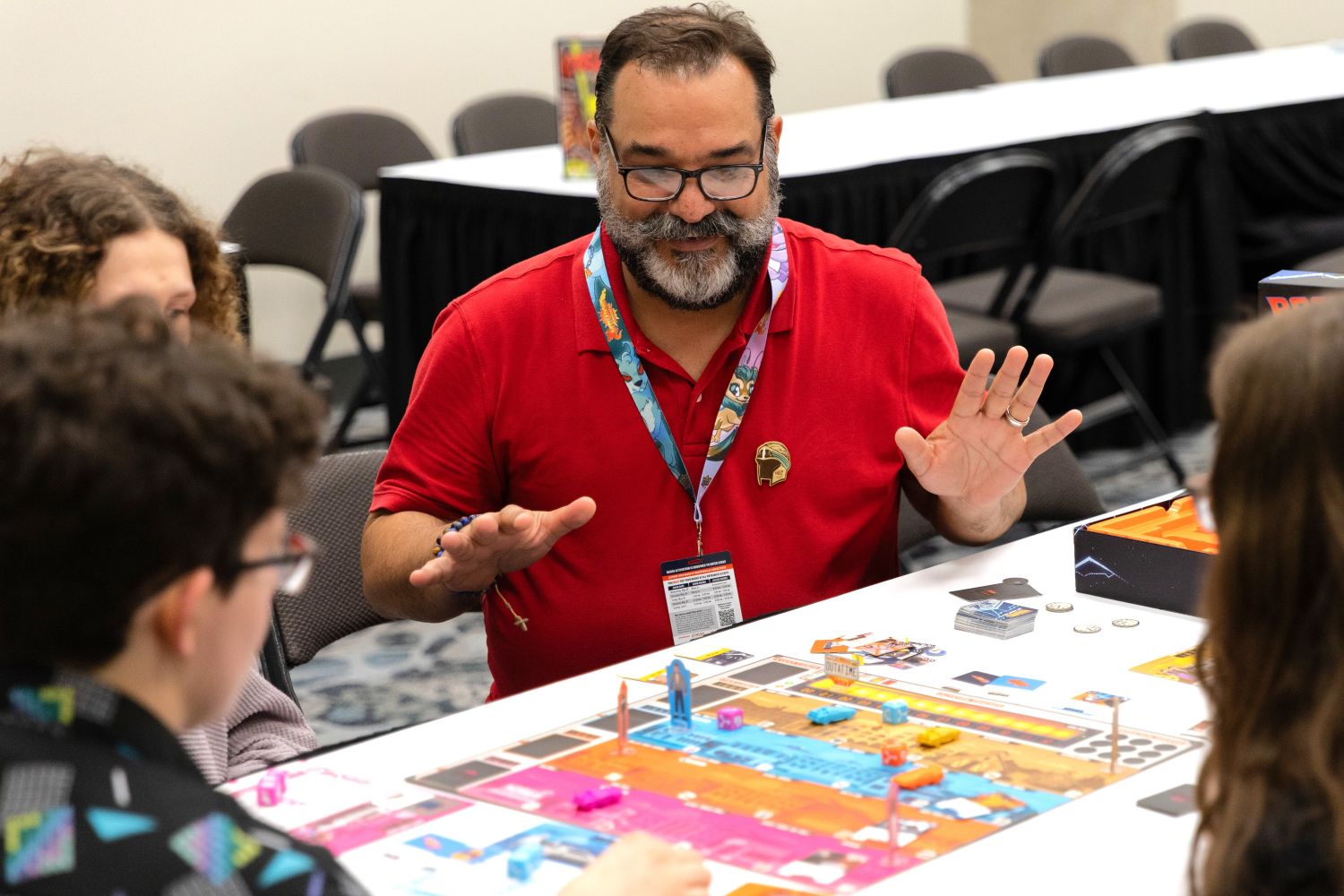 A tabletop gaming host explains rules to players gathered around a board game at Dallas FAN FESTIVAL. Brightly colored pieces and cards cover the game board as fans lean in to play.