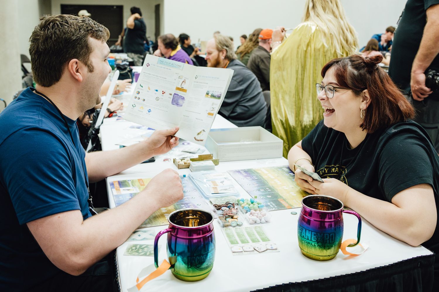 Two attendees enjoy a board game session at FAN EXPO. One holds the rulebook while the other laughs joyfully. The table is set with game pieces, rainbow metallic mugs with the FAN EXPO Dallas logo, and a colorful board game layout.