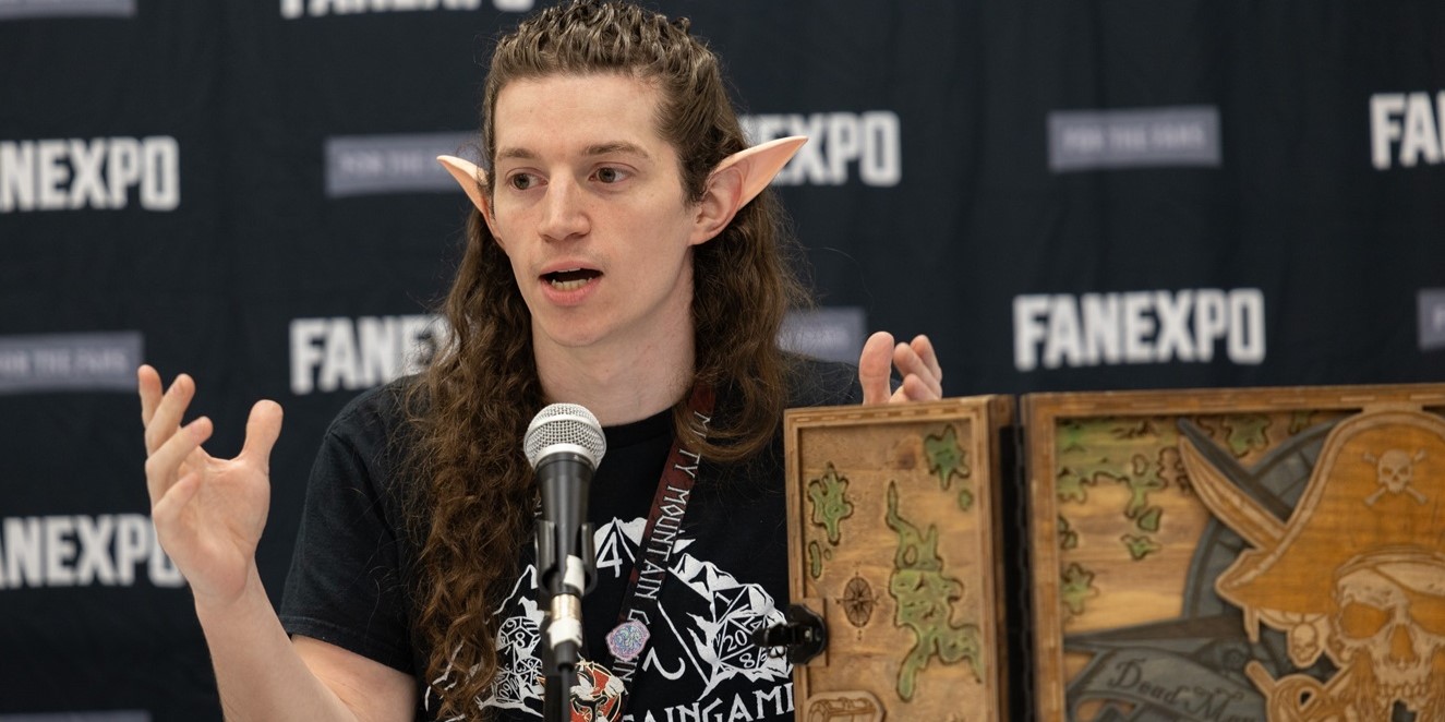 A speaker dressed as an elf with pointed ears participates in a panel discussion at FAN EXPO. They are animatedly talking into a microphone, gesturing with their hands, and sitting in front of a custom-made wooden game board featuring intricate designs. The backdrop behind them displays the FAN EXPO logo, highlighting the event's focus on gaming and fantasy.