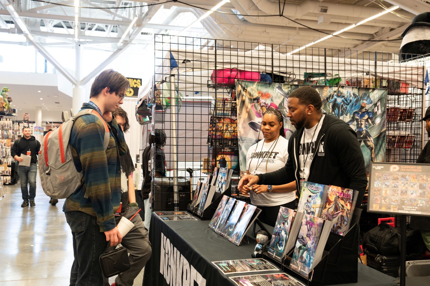 A vendor from Konkret Comics enthusiastically explains a comic title to a young man with a backpack. The booth’s display showcases vibrant indie comic covers featuring original Black superheroes. A woman in matching Konkret attire stands proudly nearby as fans explore the merchandise.
