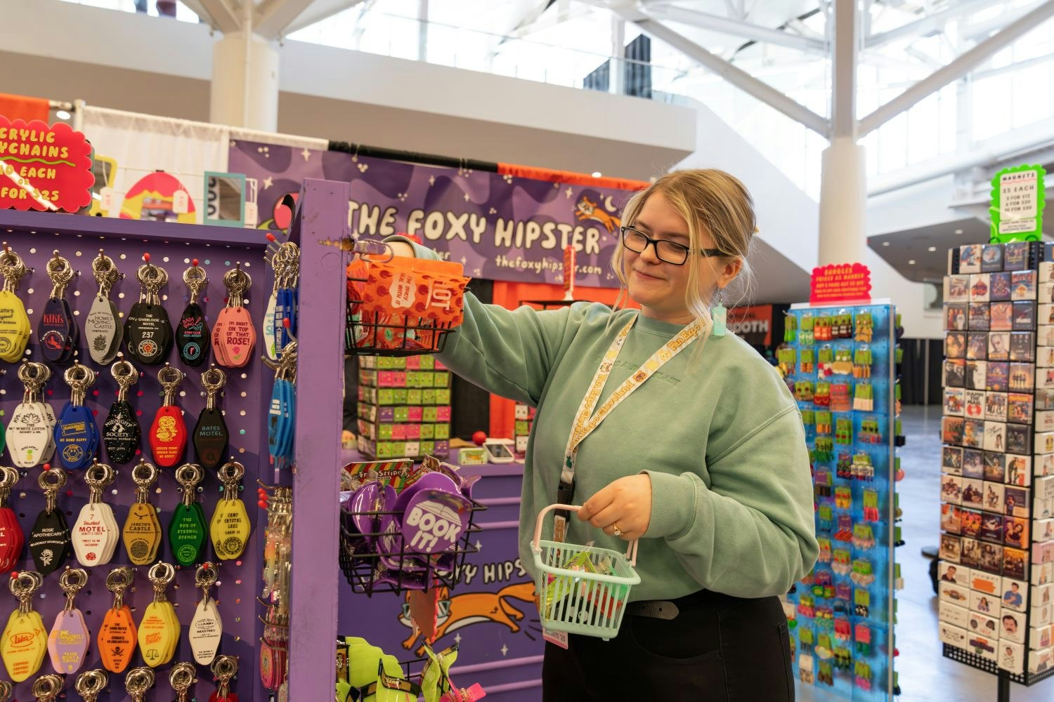 A woman in a mint green sweatshirt browses brightly colored novelty keychains and accessories at a booth called “The Foxy Hipster.” She’s smiling while holding a small shopping basket filled with fun items, standing in front of a wall of tongue-in-cheek motel key tags and quirky enamel pins.