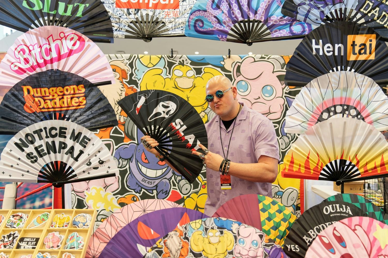 A vendor with bold fashion sense poses with a folding fan that reads “SLAY” and features the Scream ghostface. His display is surrounded by an eye-popping collage of oversized fans with cheeky phrases like “Notice Me Senpai” and “Dungeons & Daddies,” plus a cartoon backdrop featuring Pokémon characters like Jigglypuff and Psyduck.