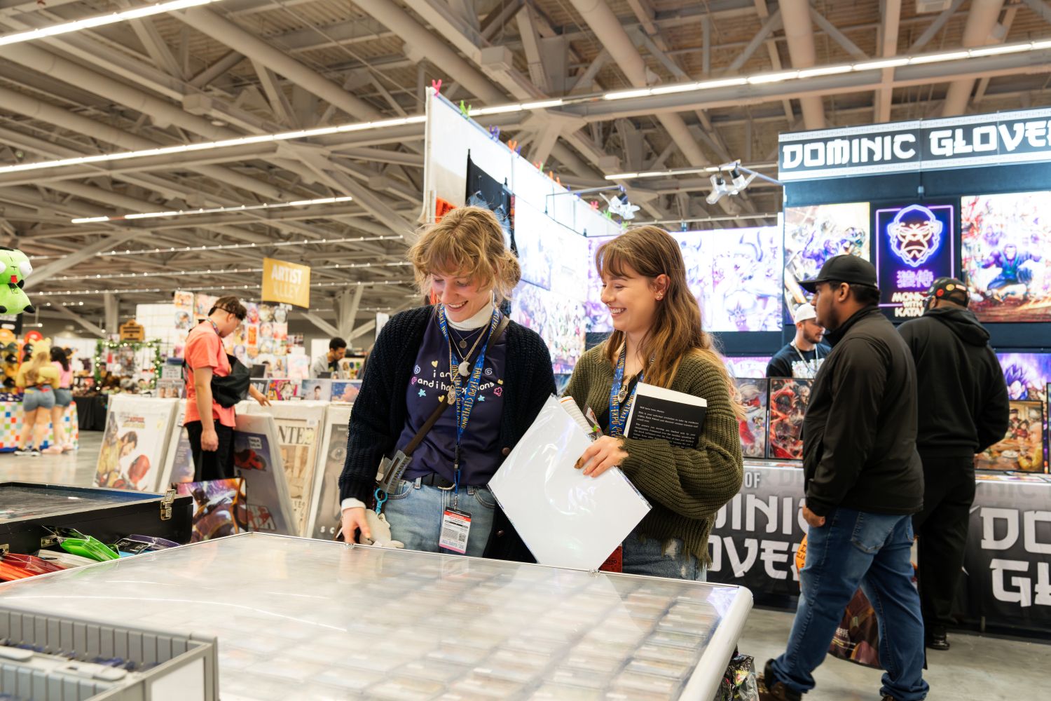 Two young women browse through trading cards at a vendor booth. Both wear FAN EXPO Cleveland lanyards and are smiling and engaged as they flip through a large binder of collectible cards. Behind them, a colorful Artist Alley booth displays fantasy and anime-style artwork under a glowing “Dominic Glover” sign.