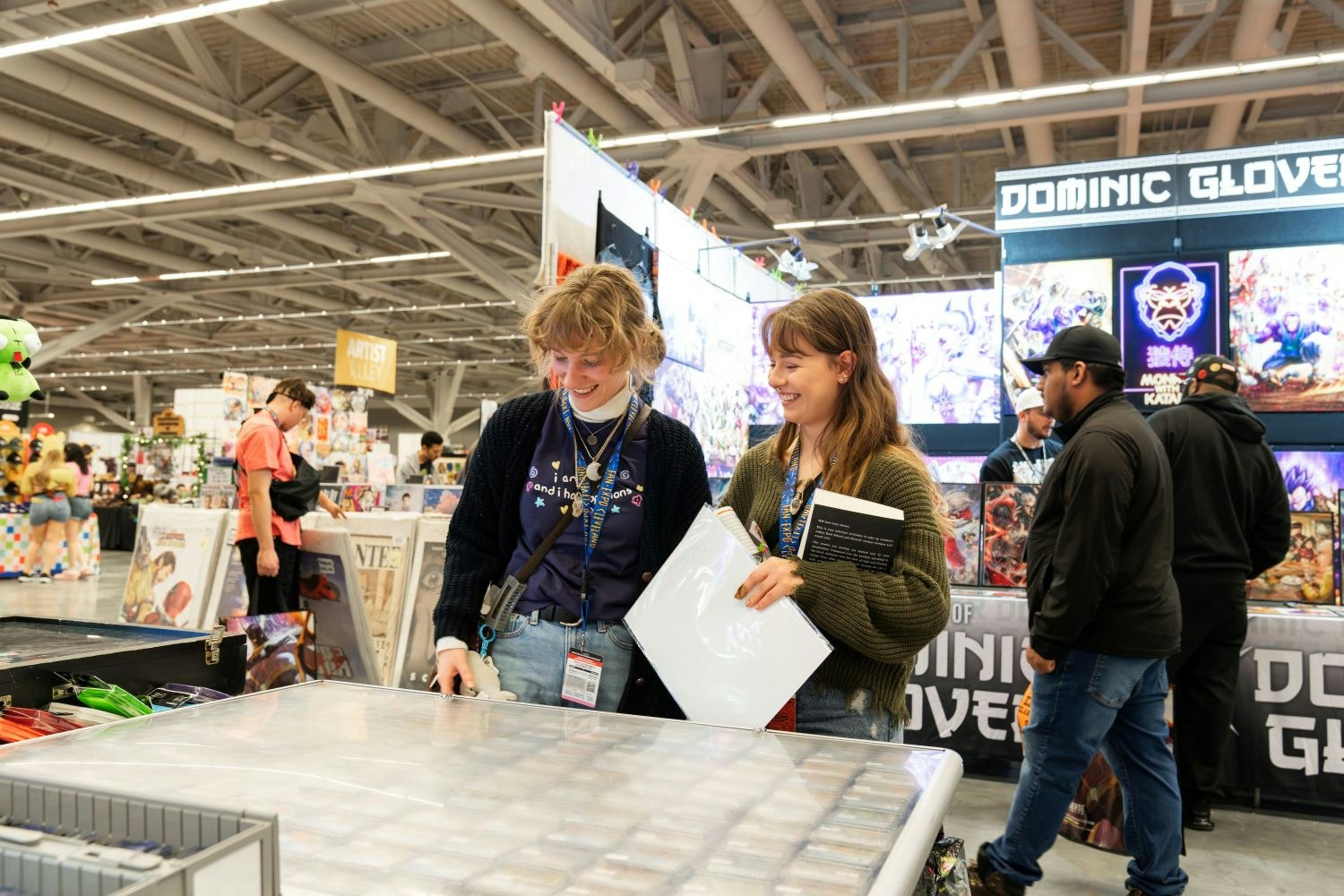 Two young women browse through trading cards at a vendor booth. Both wear FAN EXPO Cleveland lanyards and are smiling and engaged as they flip through a large binder of collectible cards. Behind them, a colorful Artist Alley booth displays fantasy and anime-style artwork under a glowing “Dominic Glover” sign.