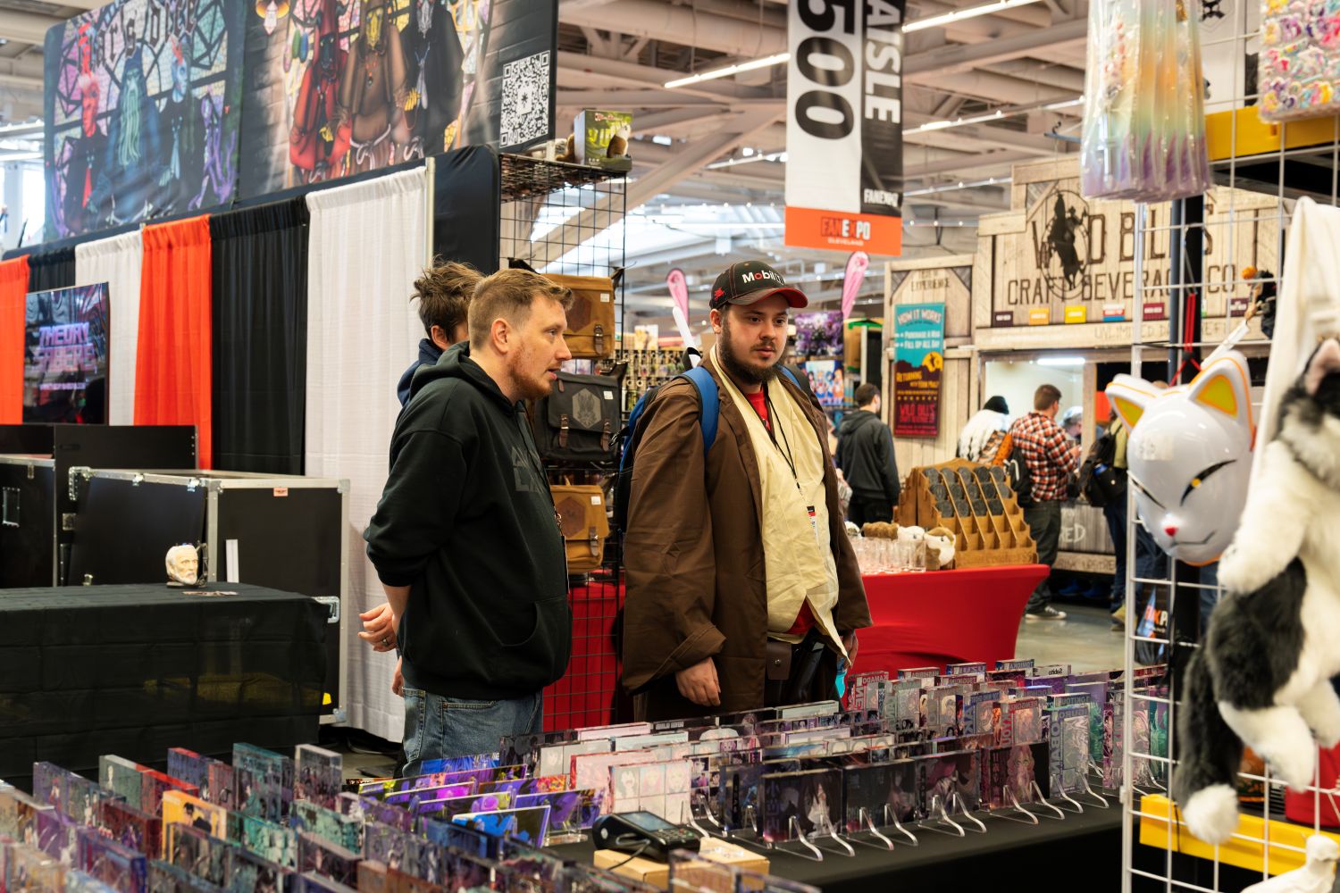 Two men examine a vendor’s display filled with anime and fantasy-themed acrylic art. One is dressed casually in a black hoodie, while the other wears a Jedi-inspired cosplay with a utility sash and a backpack. They stand next to a rack of leather goods and plush cat masks at the intersection of Aisle 500.