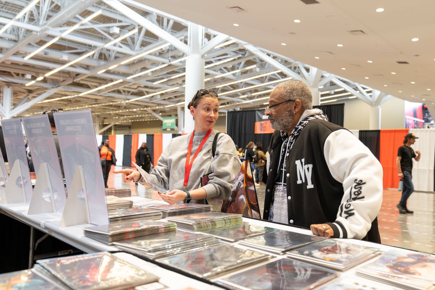 A woman enthusiastically points at a comic print while speaking with a man wearing a black-and-white NY varsity jacket. They’re standing in front of a table full of polybagged exclusives. The vibrant red lanyard and full shopping tote suggest she’s a seasoned convention-goer with a plan.