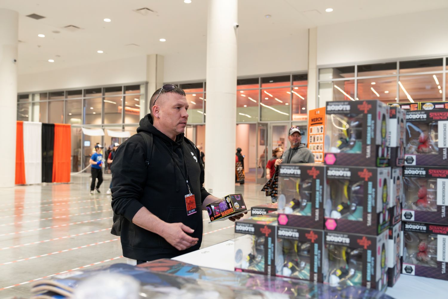 A man in a black hoodie studies boxed “Stranger Things” collectible figures stacked neatly on a table. With sunglasses resting on his head and a backpack slung over one shoulder, he clutches a few items, clearly weighing his purchase options as he shops the show floor.