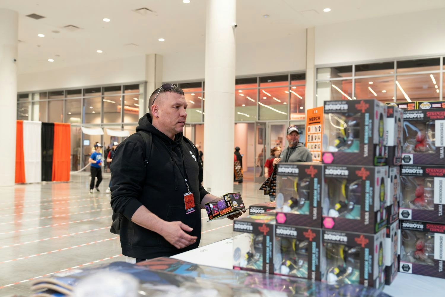 A man in a black hoodie studies boxed “Stranger Things” collectible figures stacked neatly on a table. With sunglasses resting on his head and a backpack slung over one shoulder, he clutches a few items, clearly weighing his purchase options as he shops the show floor.