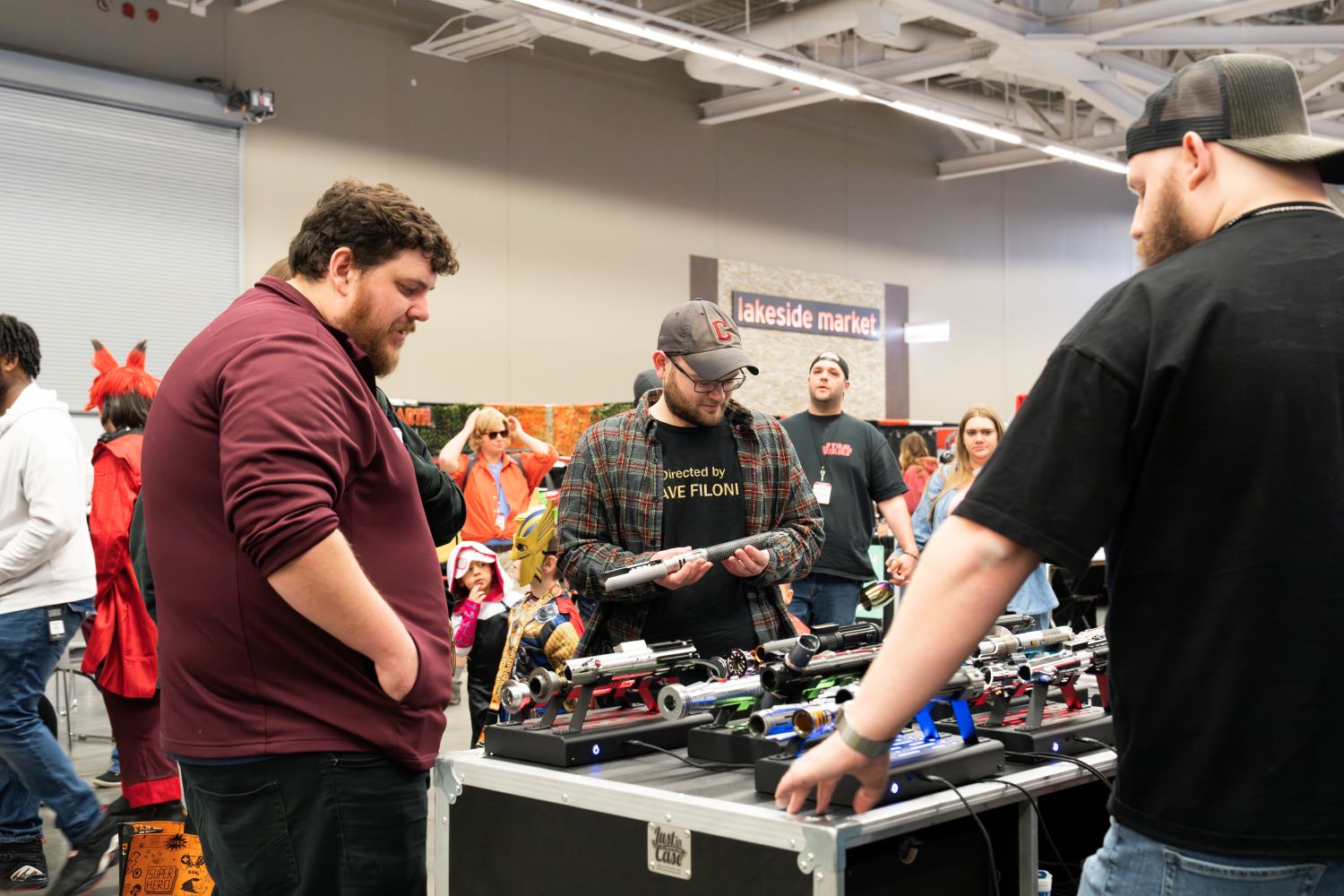 A trio of fans gather around a lightsaber booth. One inspects a hilt closely, wearing a “Directed by Dave Filoni” shirt and a Cleveland Guardians cap. A colorful array of sabers sits on display before them, while a bustling crowd passes behind the table in the background.