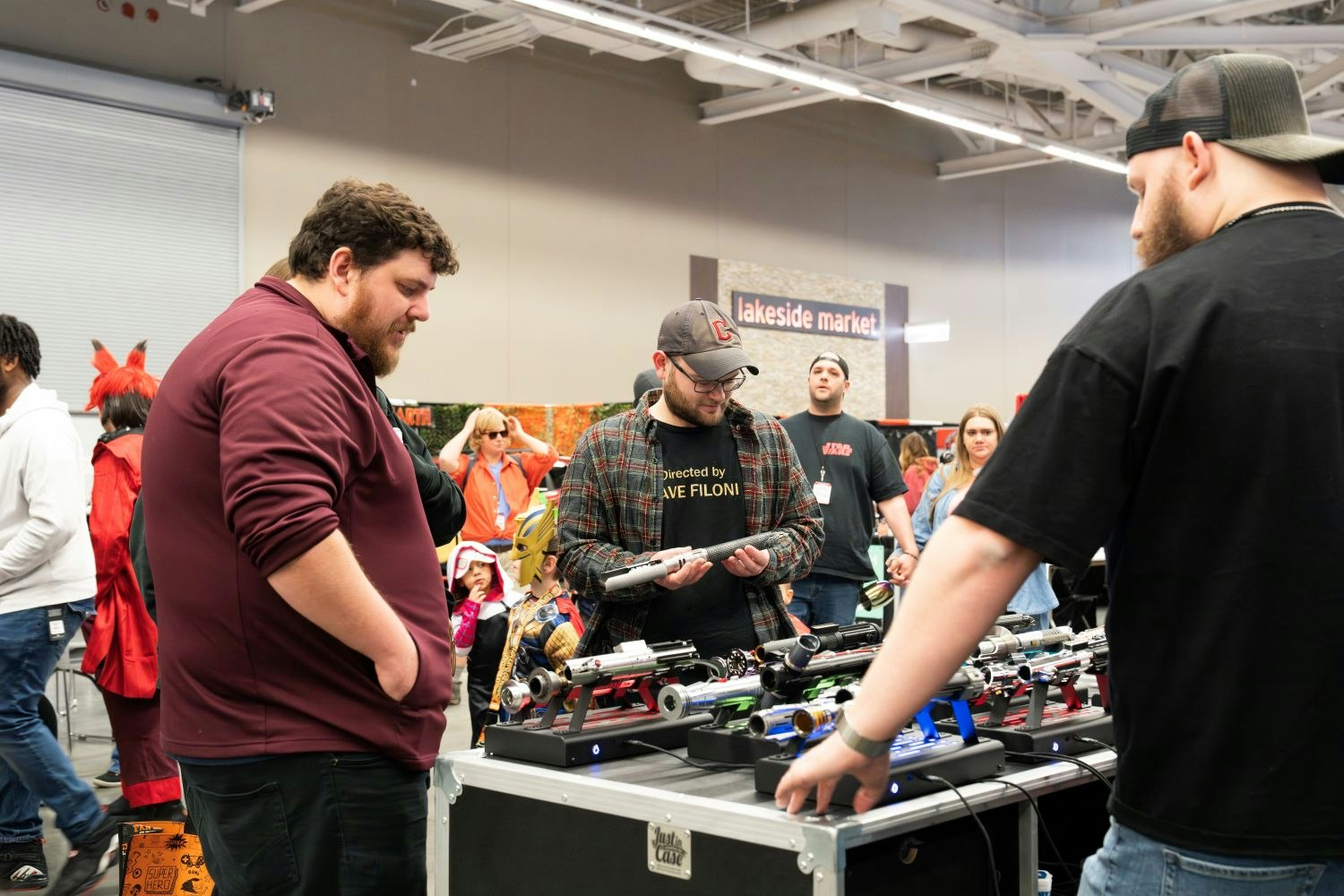 A trio of fans gather around a lightsaber booth. One inspects a hilt closely, wearing a “Directed by Dave Filoni” shirt and a Cleveland Guardians cap. A colorful array of sabers sits on display before them, while a bustling crowd passes behind the table in the background.