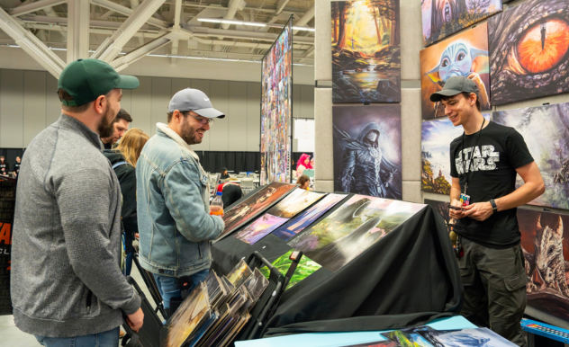 The image shows a lively interaction at a vendor booth at a FAN EXPO event. Three men, two browsing and one vendor, are engaged in conversation over a display of vibrant and intricate artwork, which includes prints of various fantasy and science fiction themes. The vendor, wearing a "Star Wars" t-shirt, is explaining details about the artworks to the two attendees, who appear interested and engaged. The booth is adorned with colorful and dramatic artworks hanging in the background, adding to the vibrant and creative atmosphere of the expo. This scene highlights the enthusiast-driven nature of such events where fans can explore and purchase unique artwork related to their interests.