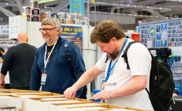 The image captures a moment at a FAN EXPO event where two men are exploring a vendor booth filled with collectibles. The man on the right is intently browsing through boxes of items, possibly comic books or other memorabilia, while the man on the left, wearing glasses and a blue jacket, looks on with a smile, enjoying the experience. The setting is a busy convention floor, with other attendees and booths visible in the background, showcasing the vibrant and bustling atmosphere of the expo. This scene highlights the communal and enthusiast nature of such events, where fans can share their passions and discover new interests.