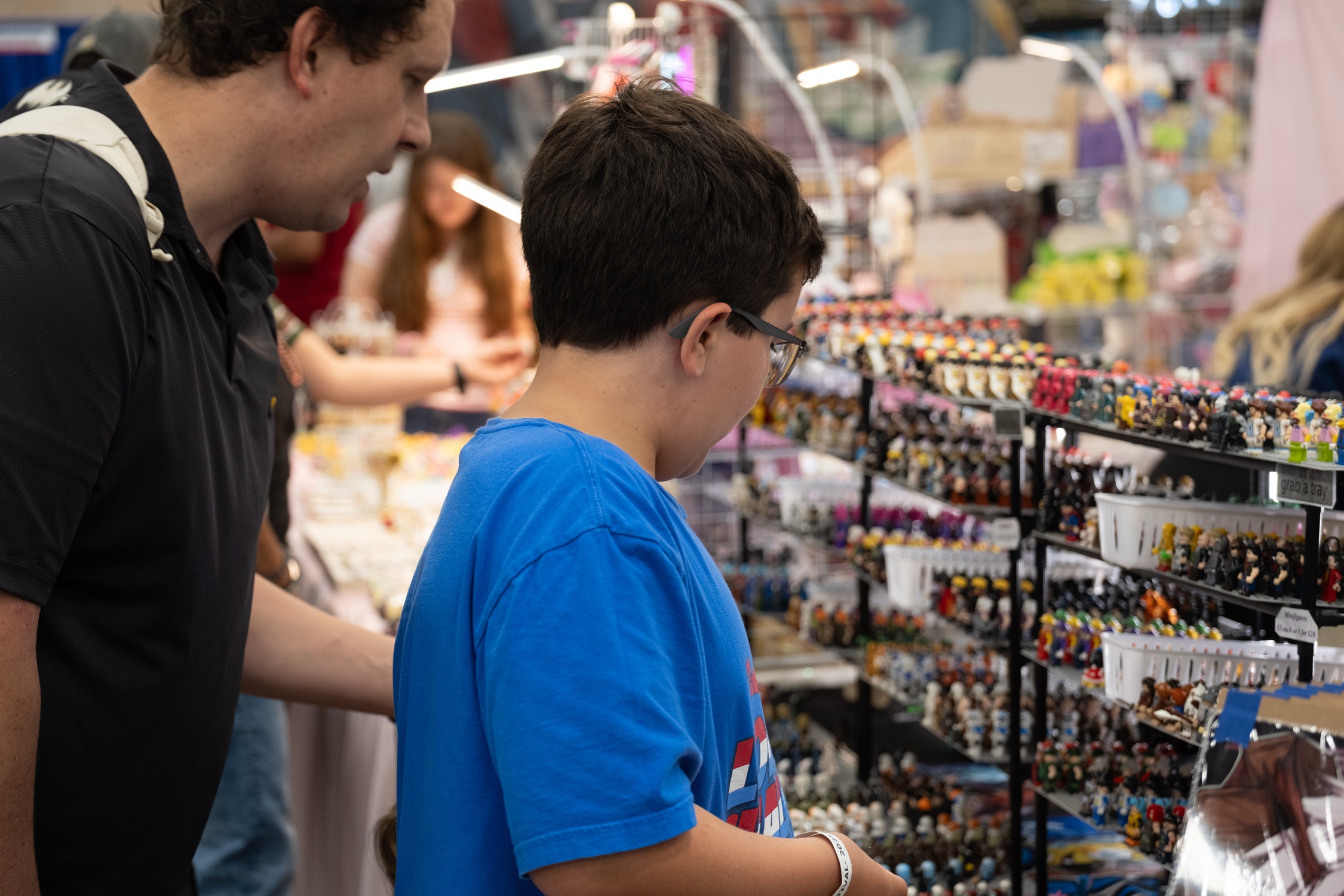 A man and young boy look closely at rows of custom mini-figures displayed on a vendor’s table, surrounded by bright lights and colorful merchandise.