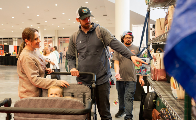 The image captures a family at a FAN EXPO event browsing through a merchandise stall. A young woman, smiling, stands beside a stroller containing a toddler, who looks curiously out. A man, presumably the father, points at an item on the shelf, possibly selecting a toy or collectible to buy. In the background, other attendees and vendors can be seen, creating a lively atmosphere typical of a fan convention. This scene showcases the family-friendly aspect of such events, where enthusiasts of all ages can enjoy exploring a wide range of pop culture merchandise together.