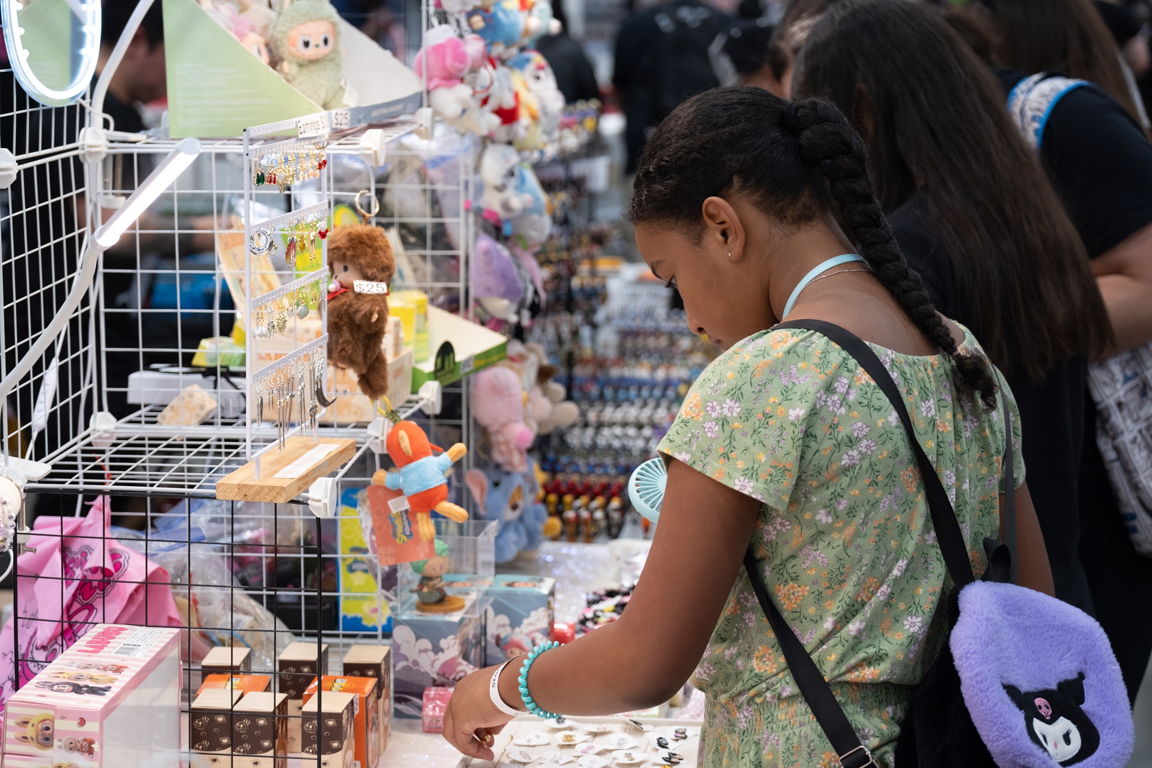 A girl in a green floral dress examines handmade jewelry and plush toys at a booth filled with cute and colorful collectibles.
