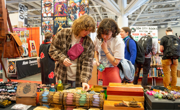The image depicts two young individuals deeply engaged in examining various collectibles at a vendor booth at a FAN EXPO event. They are looking at items such as intricately designed notebooks and colorful gemstones laid out on a rustic wooden table. The one on the left, wearing a plaid overshirt and carrying a shoulder bag, points at something interesting, while the one on the right, in a red top and holding a purse, looks on intently. The background is lively with other attendees and booths featuring comic book and pop culture merchandise, capturing the vibrant and exploratory spirit of the convention.