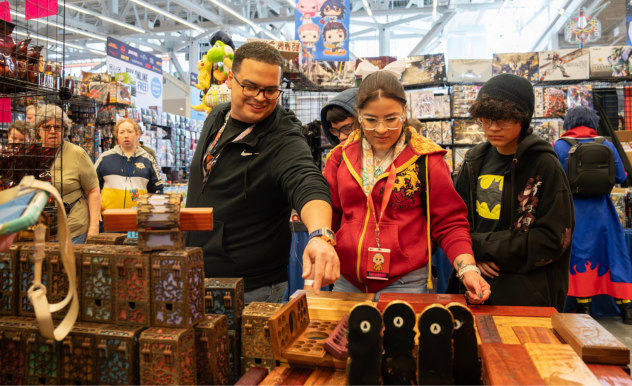 The image shows three attendees exploring a booth at a FAN EXPO event, featuring a variety of intriguing wooden boxes and collectibles on display. A man and a woman, standing close together, appear engaged as they inspect a particular item that the man is pointing at, while a younger individual, wearing a Batman hoodie, looks on with interest. The booth is packed with assorted pop culture merchandise, from plush toys to decorative items, capturing the diverse interests of the attendees. The scene is lively, with other shoppers and booths visible in the background, illustrating the vibrant atmosphere of the expo where fans gather to celebrate and indulge in their favorite fandoms.
