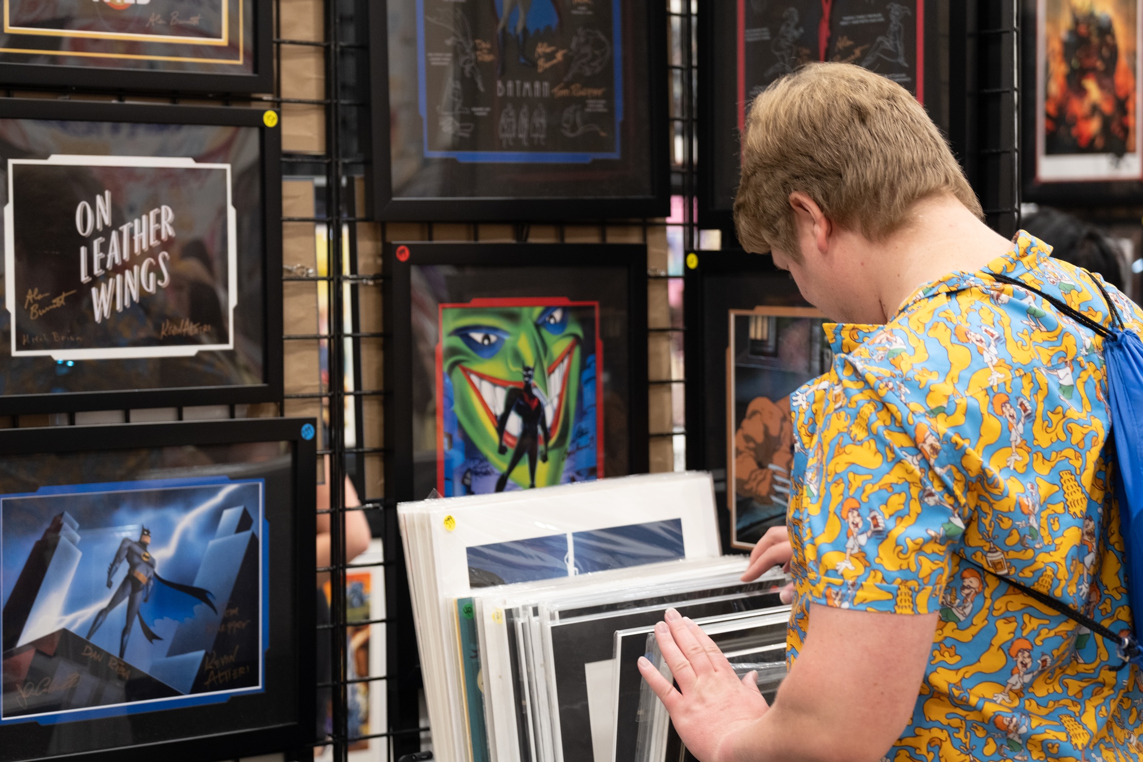 A fan in a patterned shirt flips through framed artwork featuring Batman: The Animated Series prints and comic-style pieces at an art vendor’s booth.
