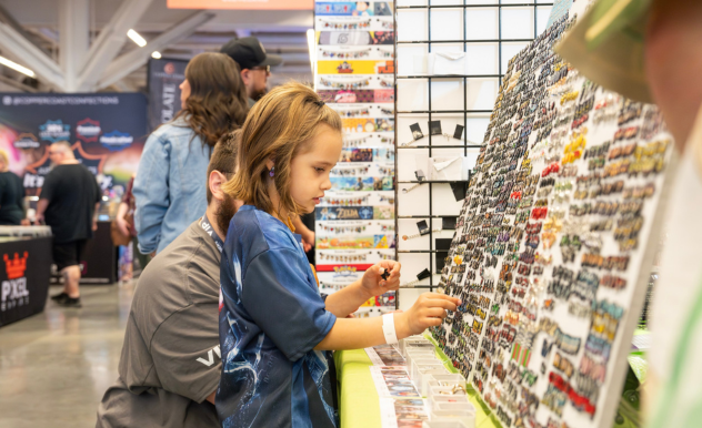 The image captures a young girl at a FAN EXPO event, intently browsing through a collection of colorful enamel pins displayed on a stand. She appears focused as she carefully examines the variety, likely selecting her favorite characters or designs. Behind her, an adult, possibly a parent, is supervising her while also engaged in looking at other merchandise. The background shows a bustling convention atmosphere with other attendees and various booths, reflecting the vibrant and family-friendly environment of the event where enthusiasts of all ages can explore and enjoy their passions.