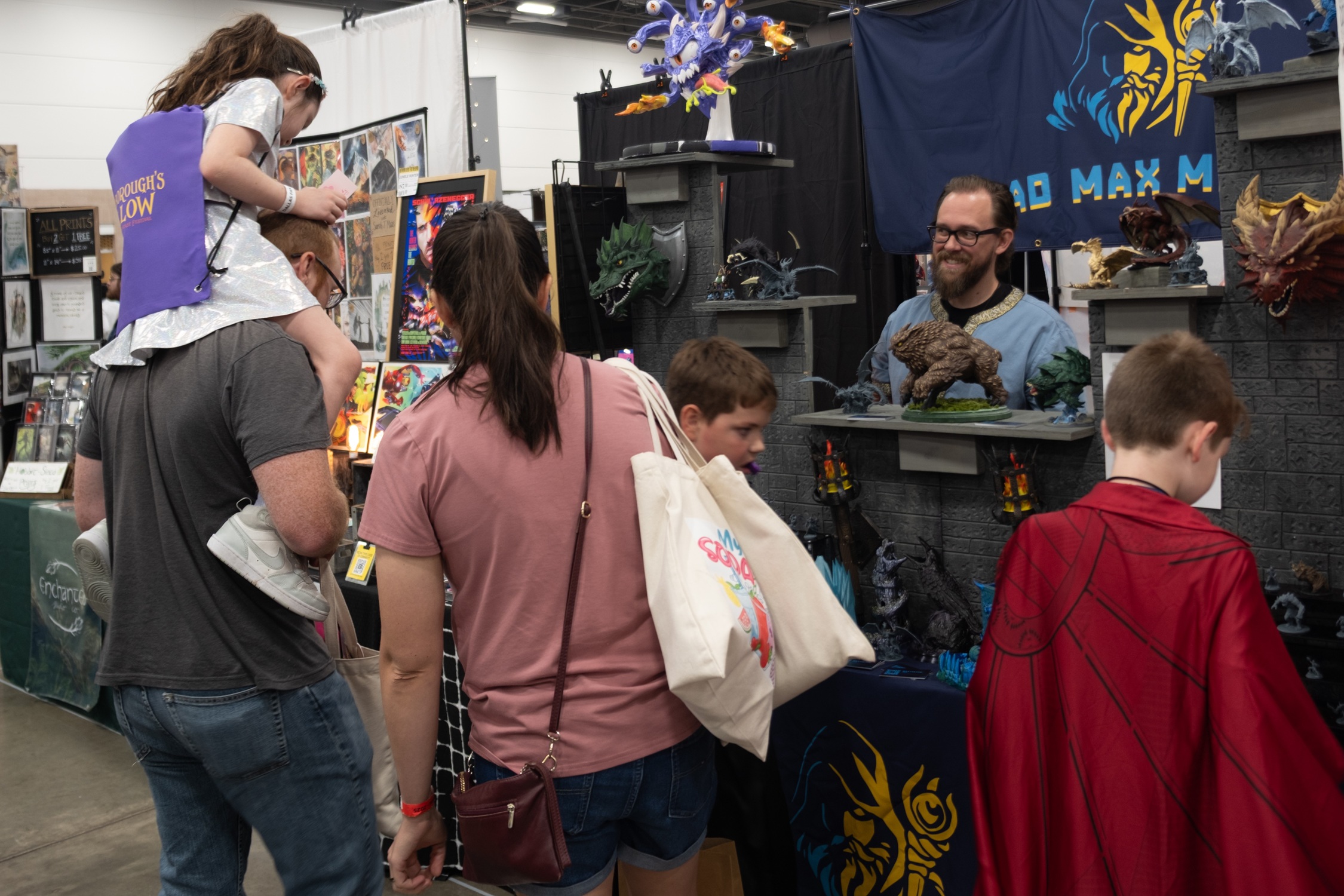 A family explores a vendor booth showcasing fantasy creature sculptures and dragon art, with a young girl perched on her dad’s shoulders.