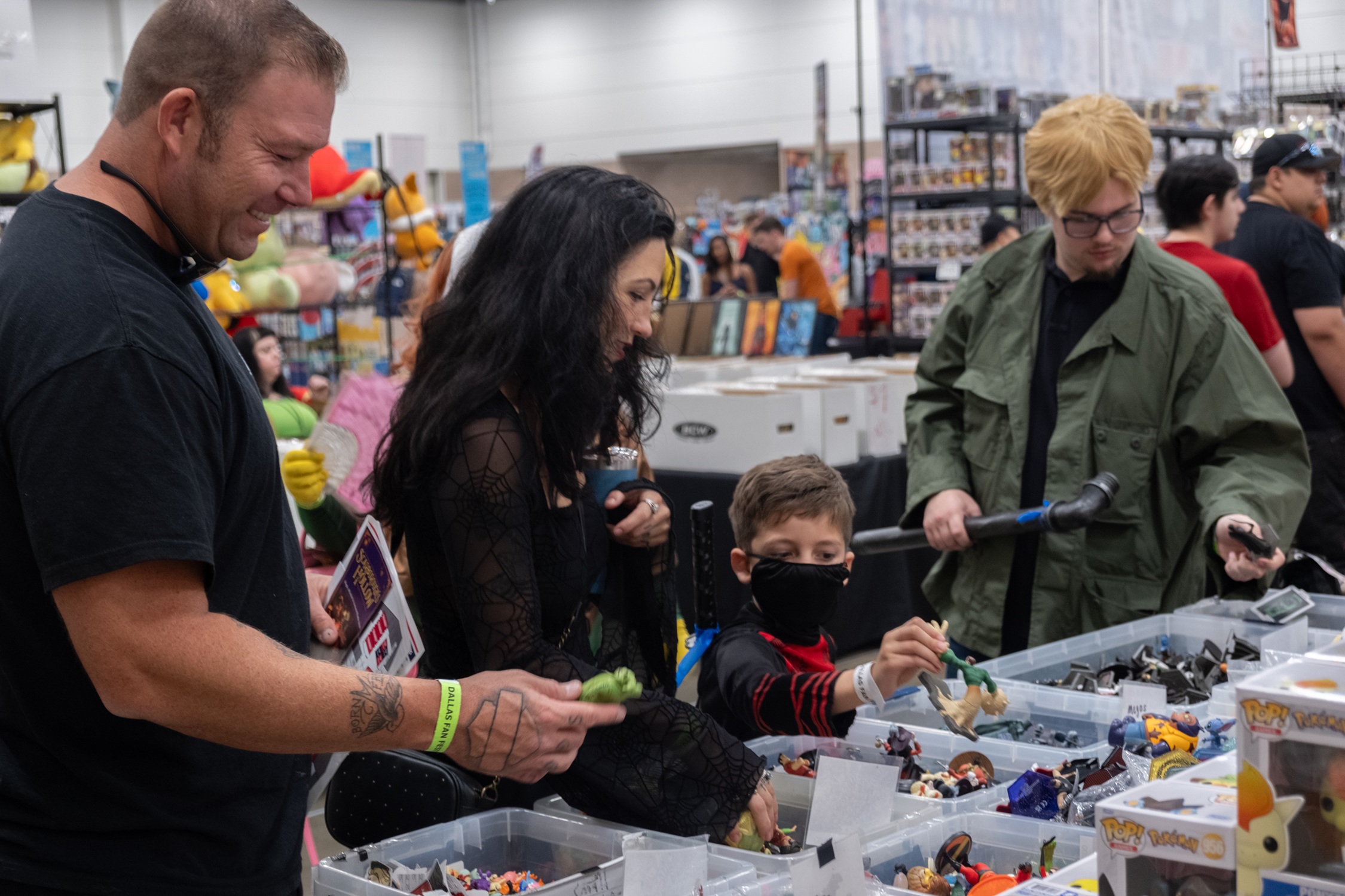 A family dressed in casual cosplay browses bins of loose action figures and collectibles, smiling and comparing their finds.