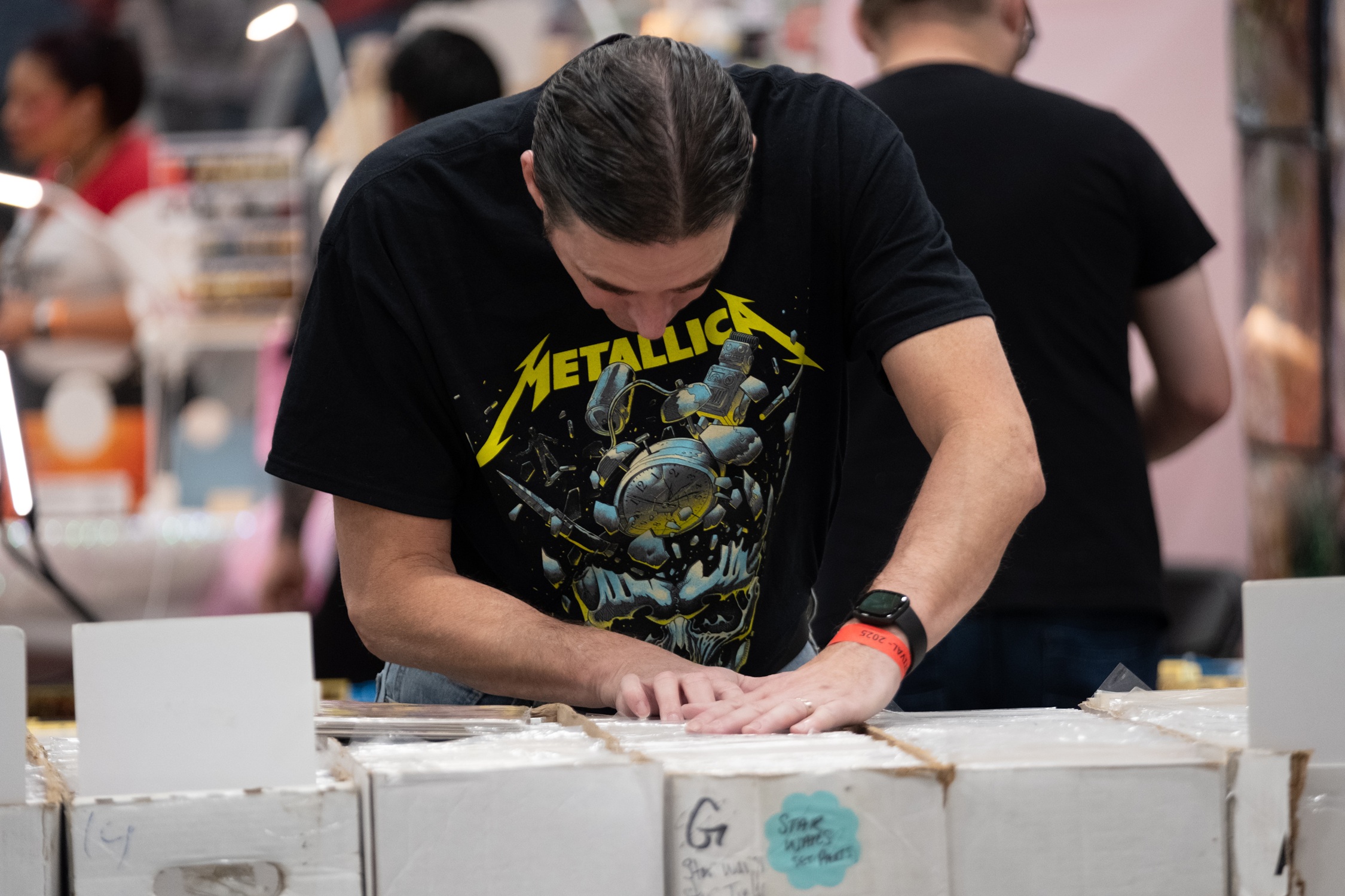 A fan in a Metallica shirt flips through long boxes of comic books, searching for new additions to their collection.