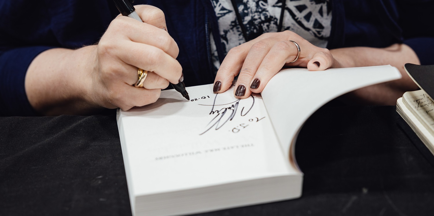 Close-up of a person signing a comic book with a black marker at an autograph booth. Fans eagerly await as the artist personalizes the signature, creating a memorable moment for collectors.