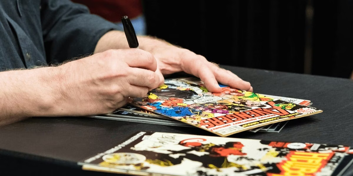 A close-up of a comic book being autographed, featuring vibrant Avengers cover art.