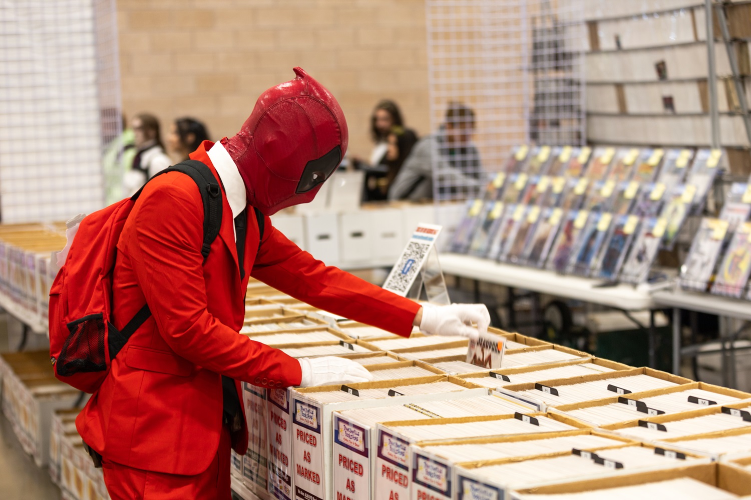 A cosplayer dressed as Deadpool with white gloves browses through boxes of comic books at FAN EXPO, closely examining a comic.
