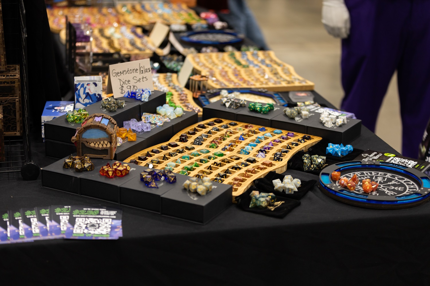 Close-up of a table at FAN EXPO displaying a colorful assortment of gemstone and glass dice sets arranged in rows. A sign on the table reads "Gemstone Glass Dice Sets $30."