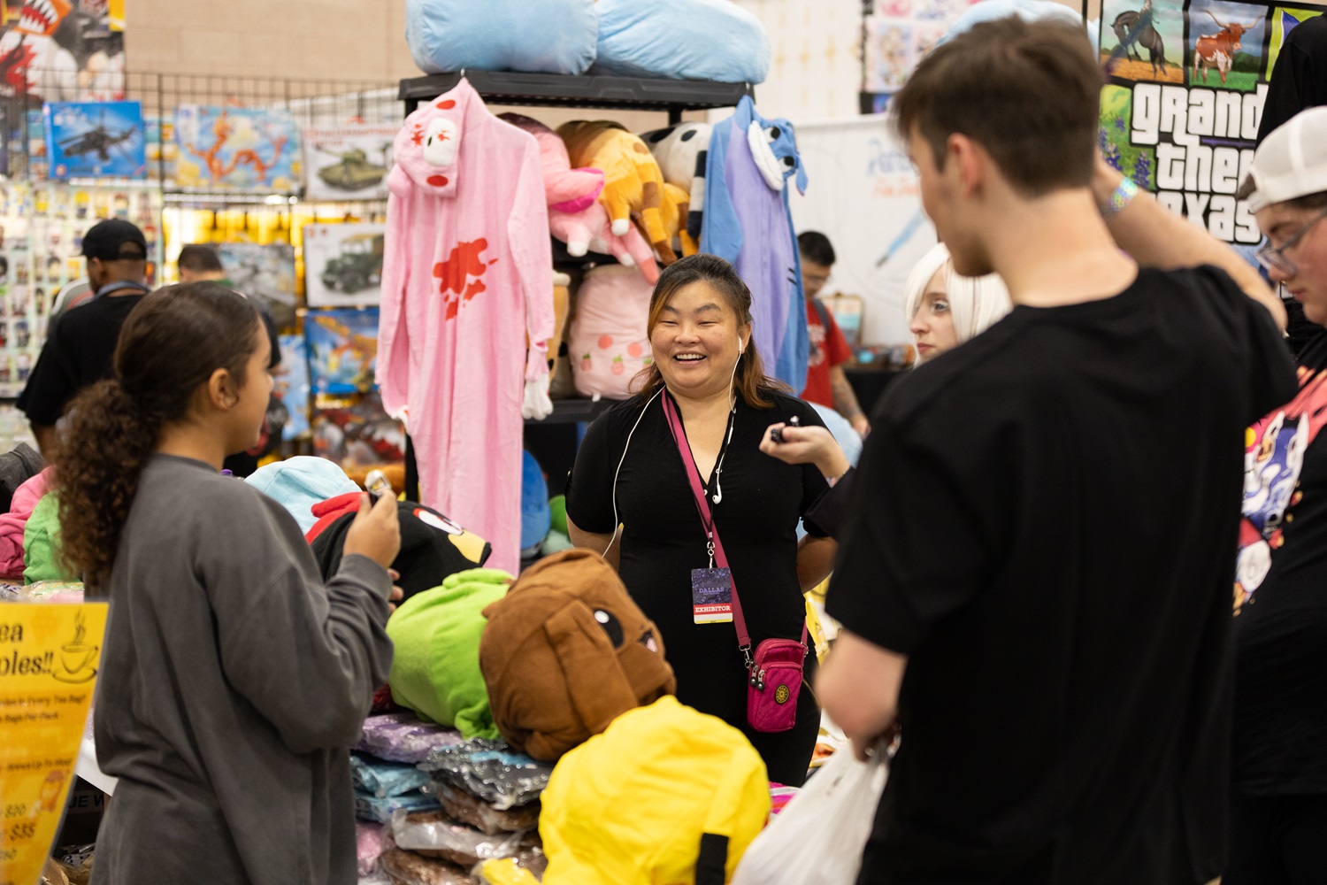 A vendor interacts with a group of attendees at a booth filled with plush toys and colorful onesies at FAN EXPO. The booth is busy with various plush options.
