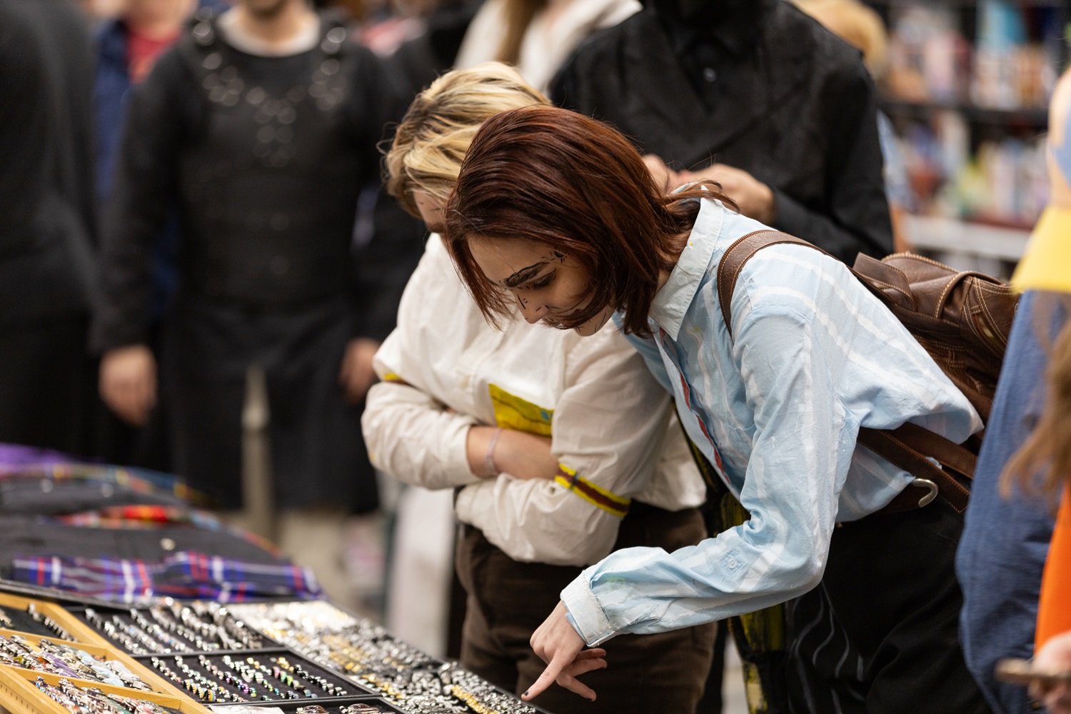 A cosplayer and another attendee look closely at a display of jewelry at FAN EXPO, pointing out specific items as they browse.